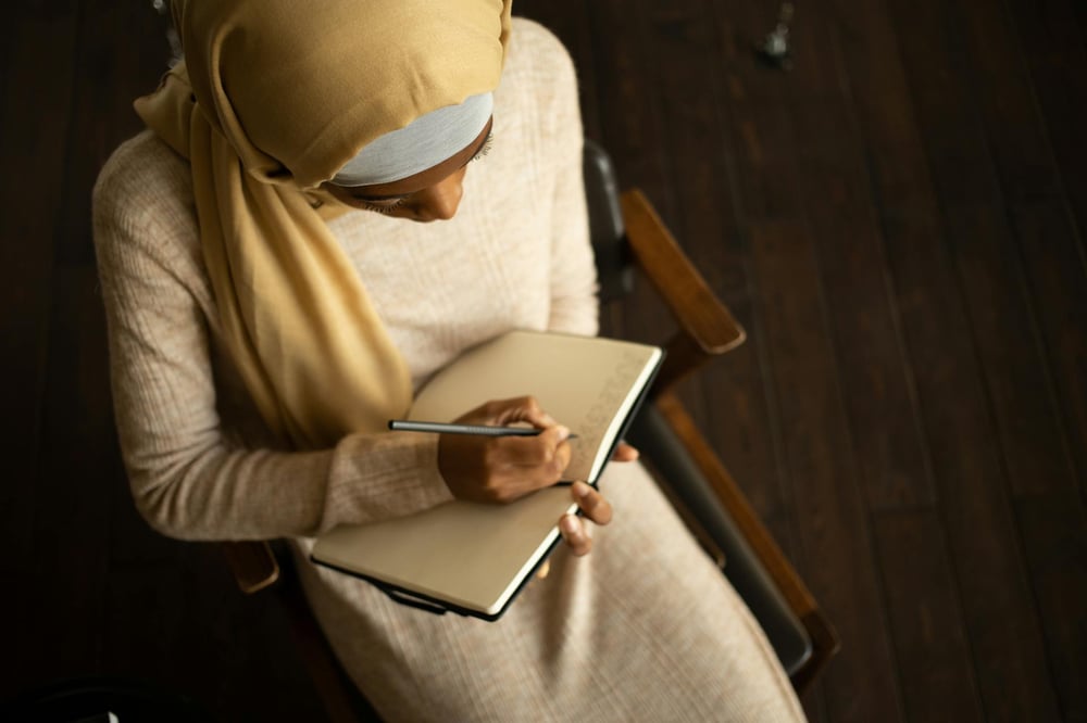 A writer sits in a wooden chair and writes in a notebook.