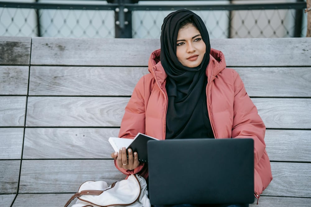 A person sits on a bench holding a laptop and an open notebook, staring thoughtfully into the distance.