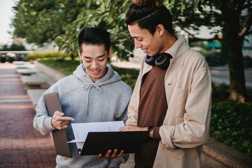 Two people stand together in a park, discussing something written on a sheet of paper.