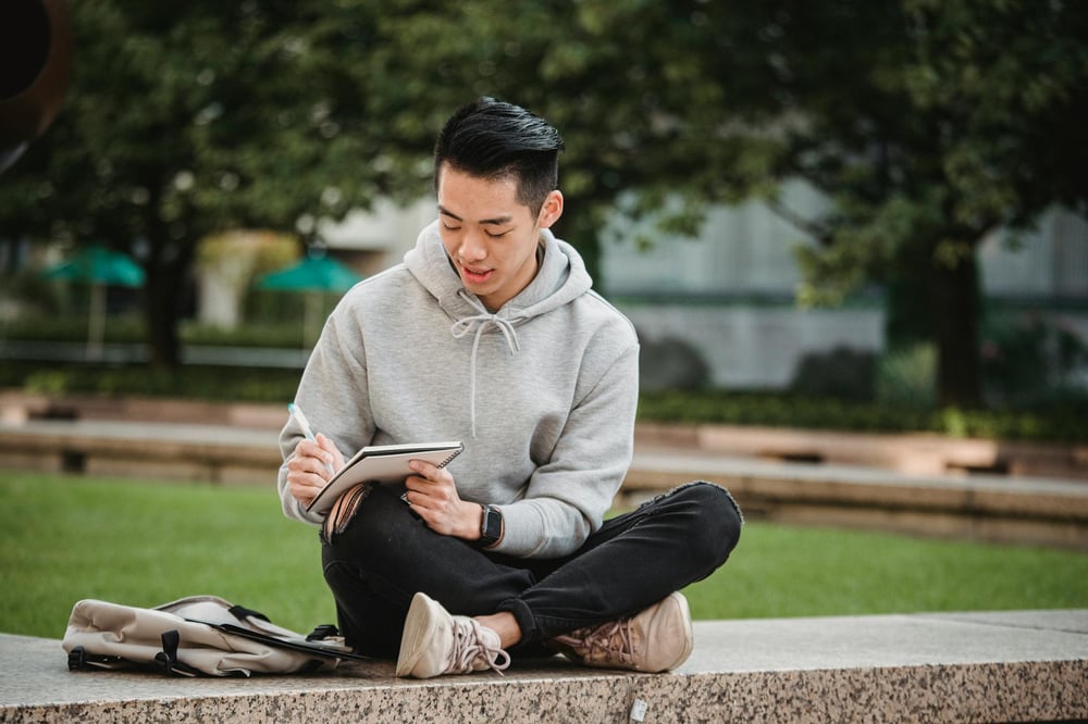 A person sits cross-legged on a low marble wall in a park, writing in a notebook.