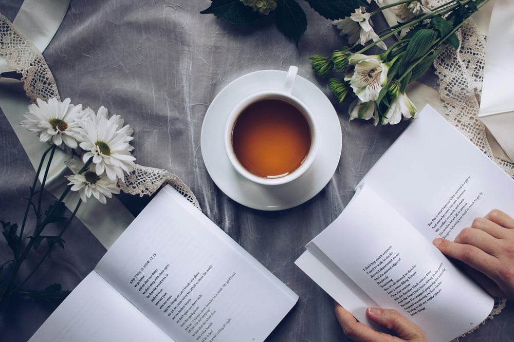 A hand holds a book of poetry open beside another open book of poetry, a white teacup, and white flowers all on a delicate white tablecloth.l