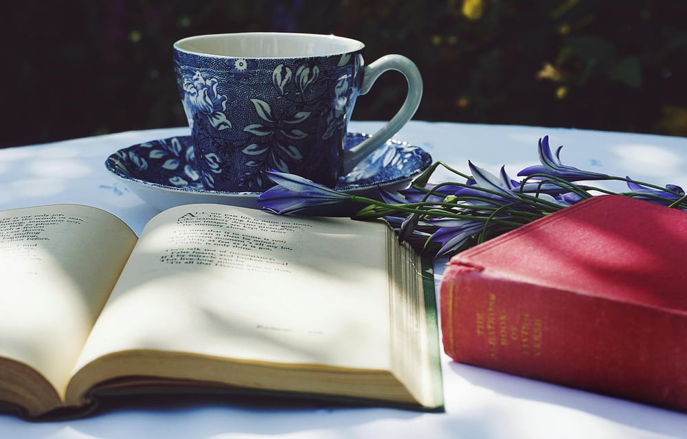 A book of poetry lays open on a white table beside a closed red book, purple flowers, and a blue and white teacup.