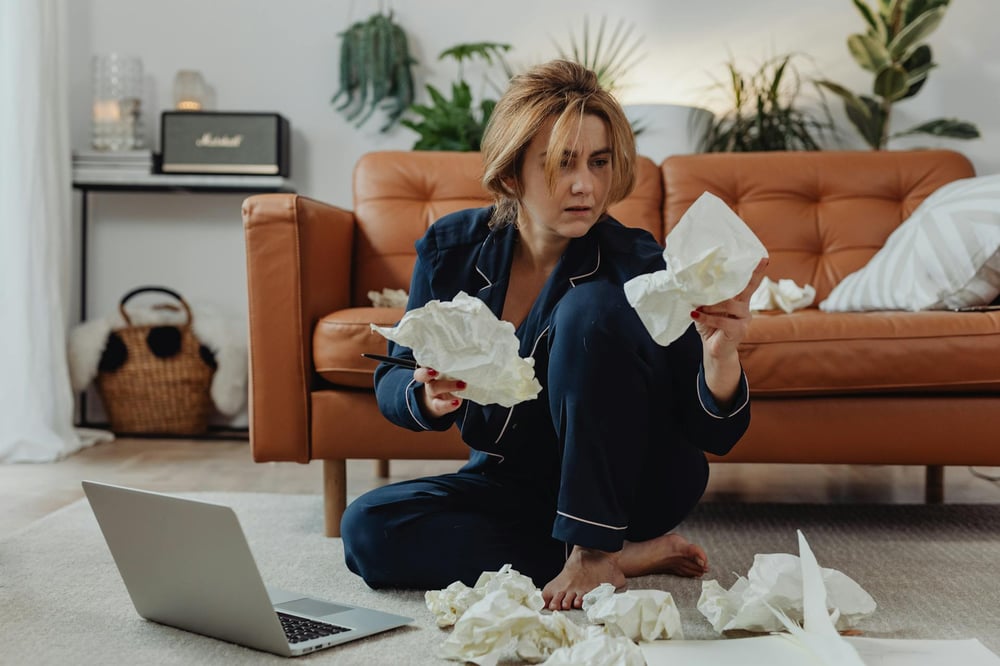 A writer wearing blue pajamas sits on the living room floor with a laptop, staring at crumpled papers.