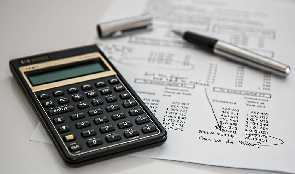 A calculator and silver pen sit on top of an annotated budget sheet.