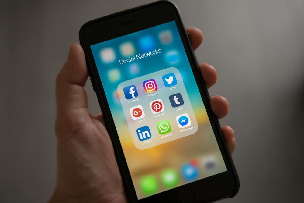 A hand holds a smartphone with icons for different social media platforms on it.