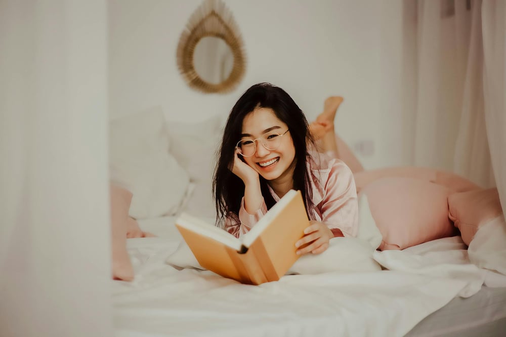 A person wearing pink pajamas and round glasses lies on a white bed reading a book.