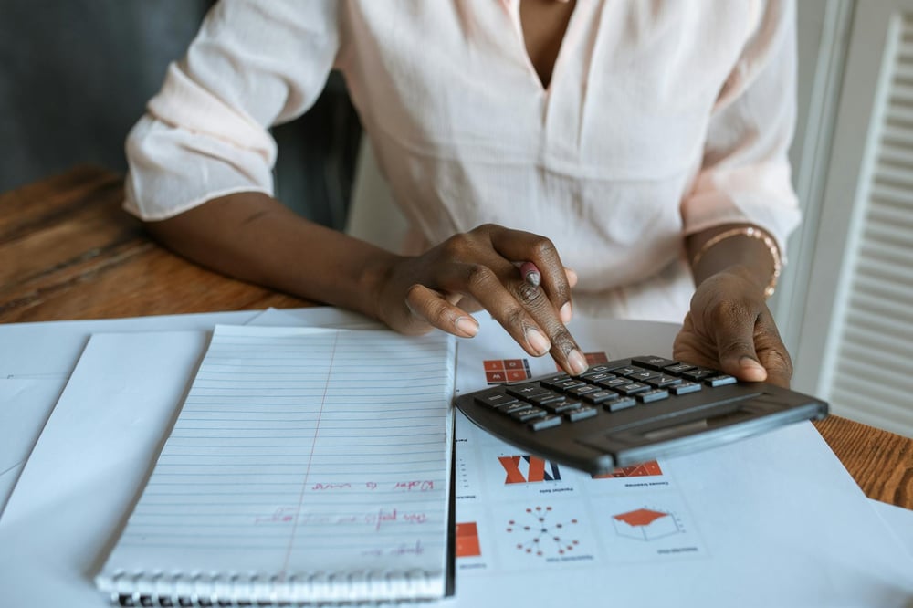 A person punches numbers into a calculator while sitting at a desk covered in papers and a notebook.