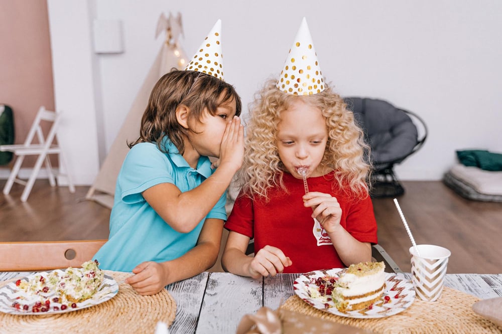 A child wearing a party hat whispers to another child who's eating cake.