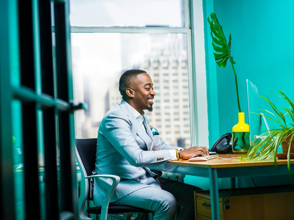 A smiling person in a blue suit types on a computer in a blue office.