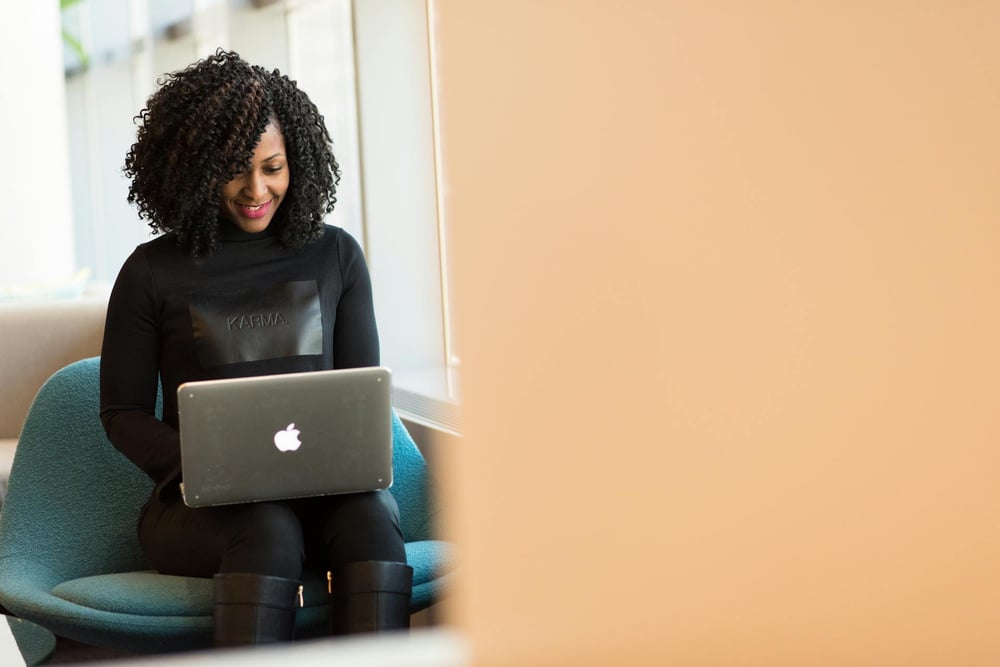 A peson sits in a blue chair by a window, working on a laptop.