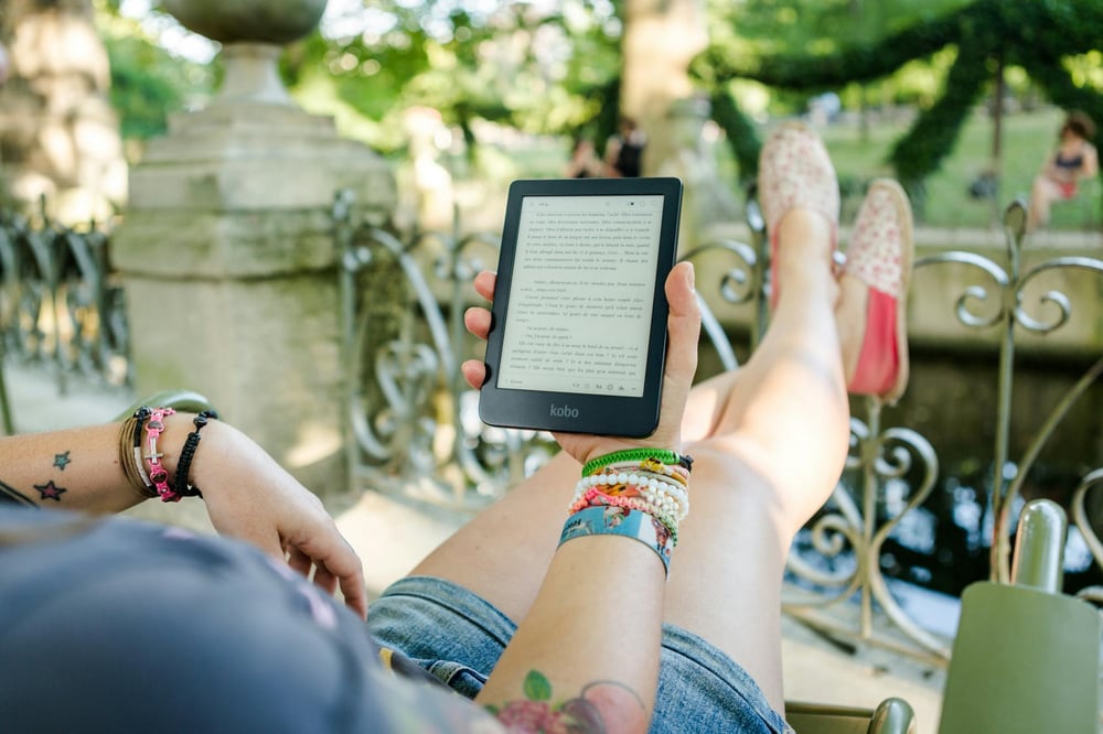 A person with jean shorts, colorful tattoos and a ton of bracelets sits on a chair outside, reading an e-reader.