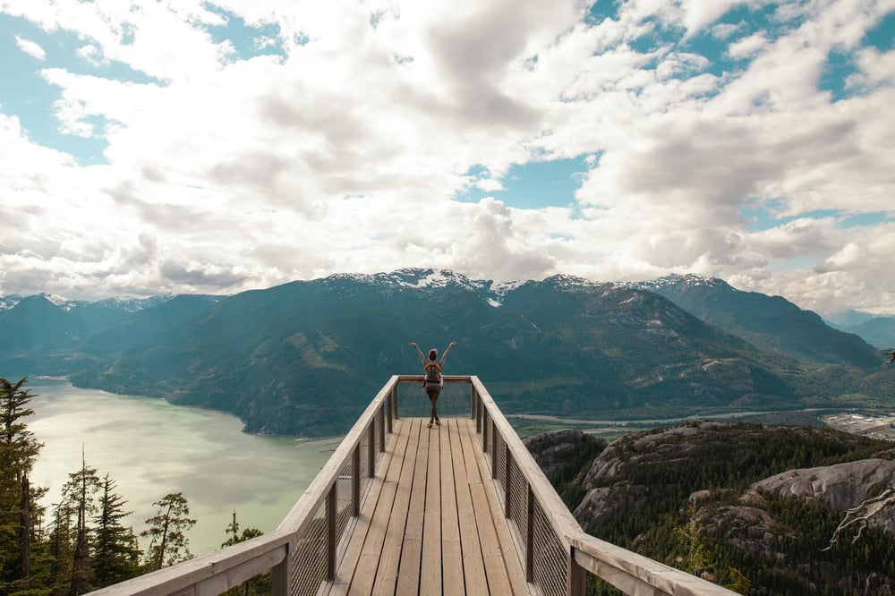 A person stands at the edge of platform that juts out over a mountain lake.