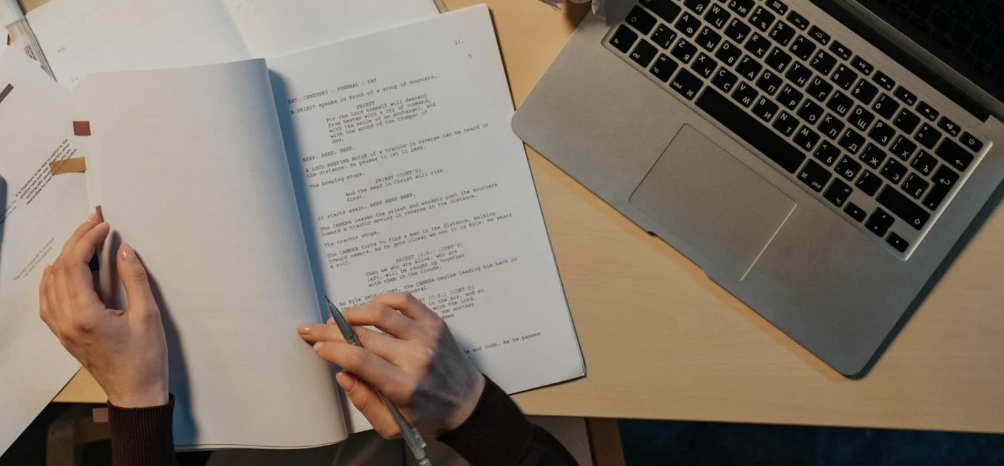 Hands hold a screenplay open on a desk beside a laptop.