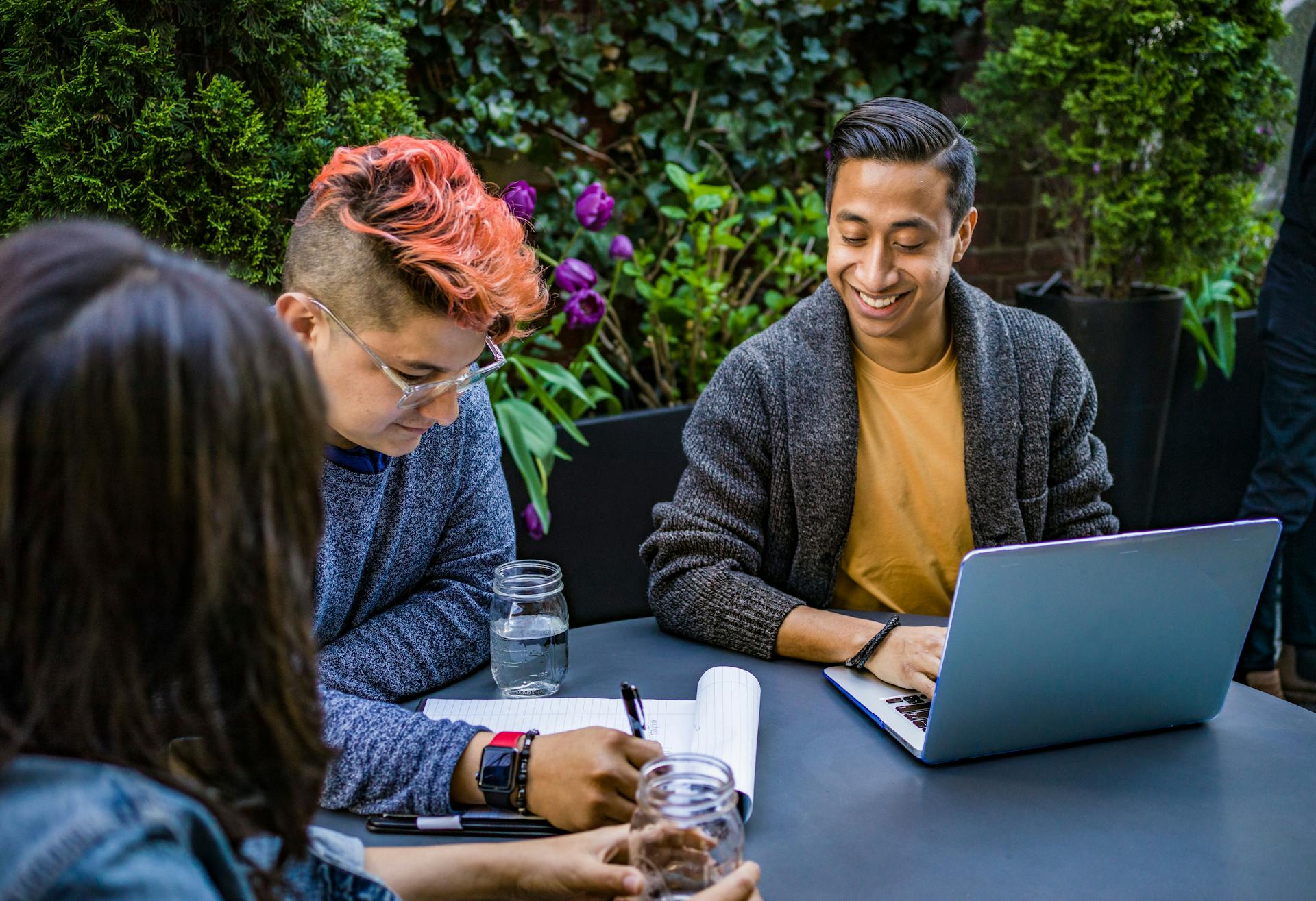 Three smiling people sit at an outdoor table writing on notepads and typing on a laptop.