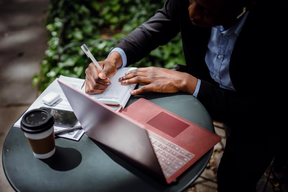 A person sits in front of an open laptop at an outdoor bistro table, writing in a notebook.