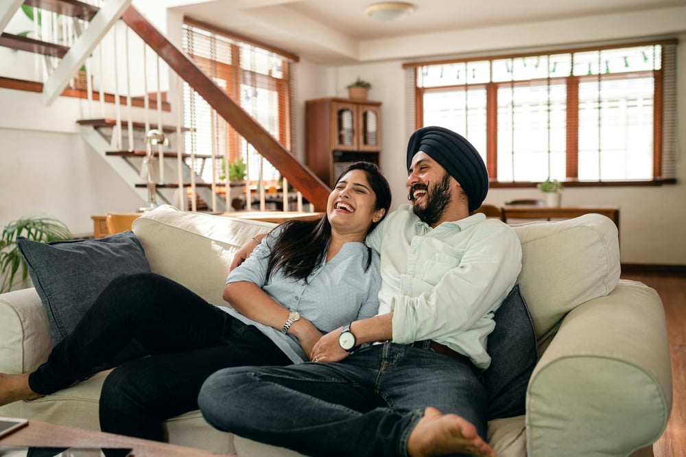 A happy couple sits together on he couch, laughing at a TV screen.