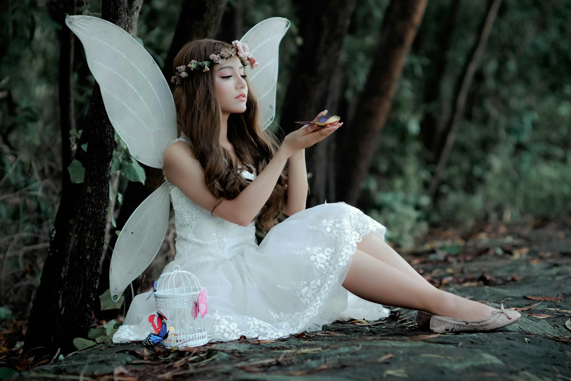 A person dressed as a fairy in a white dress and white wings sits at the foot of a tree, holding a butterfly.