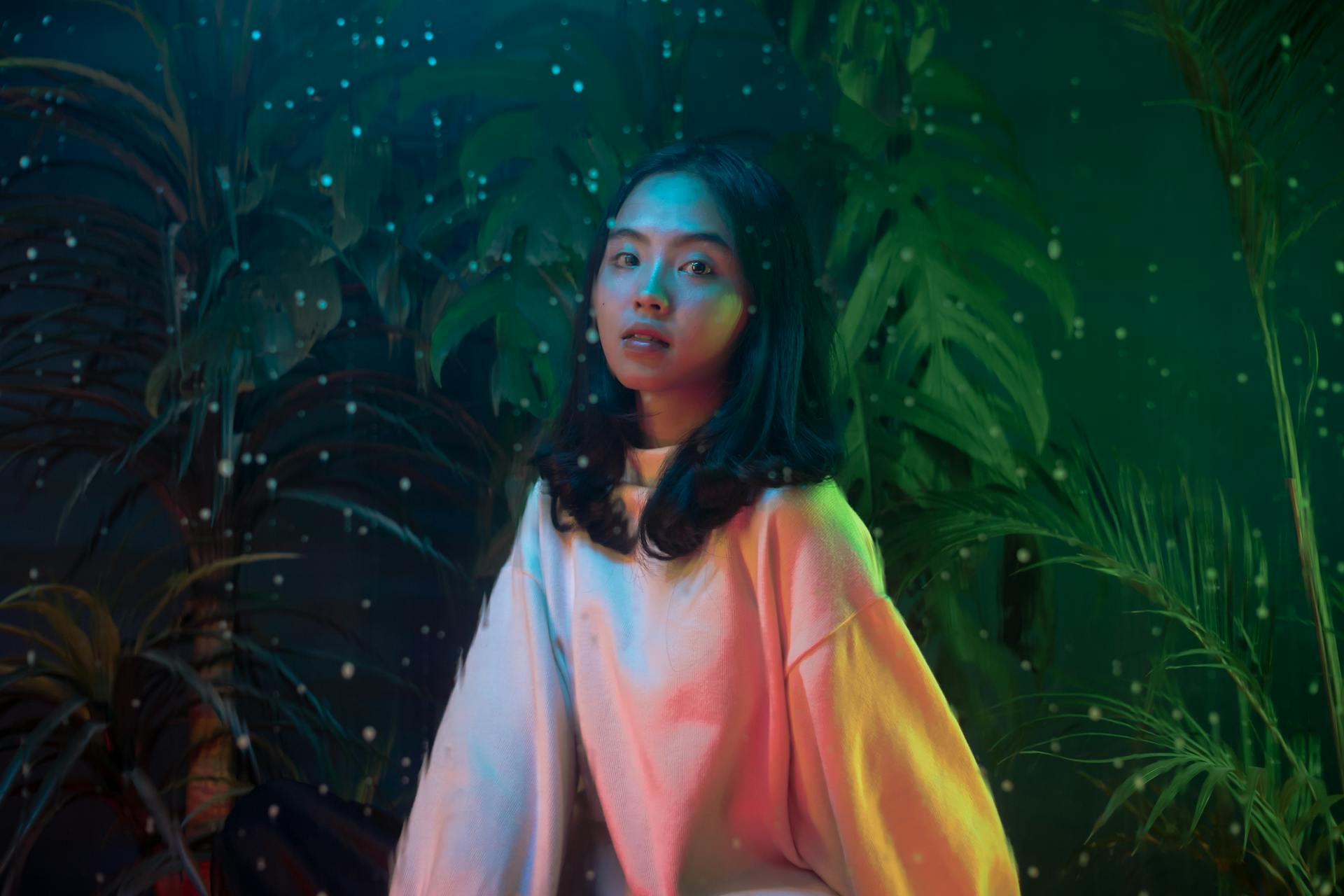 A person in a sweatshirt stands in front of tropical plants, surrounded by little dots of light.