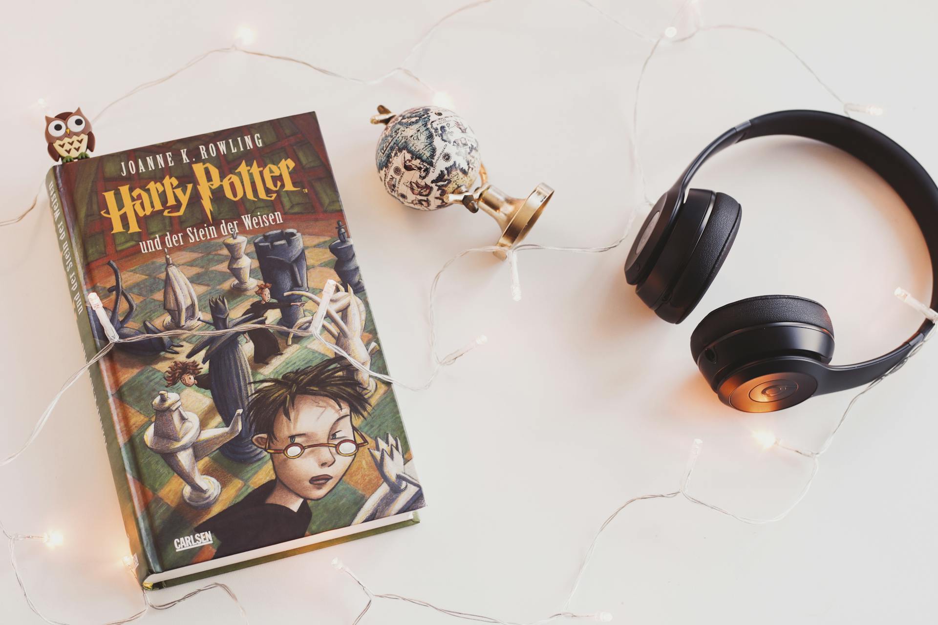 A copy of Harry Potter on a white surface beside a small globe, headphones, and string lights.