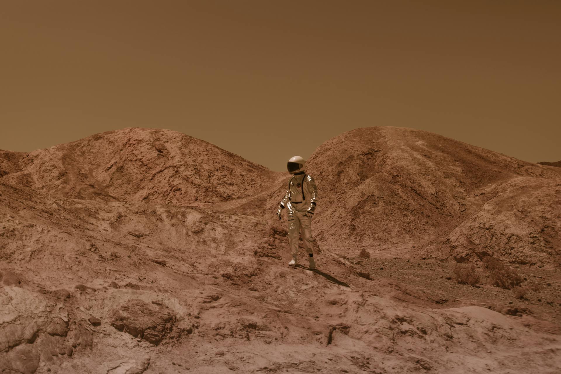 A person in a silver spacesuit standing on a dry, hilly planet.