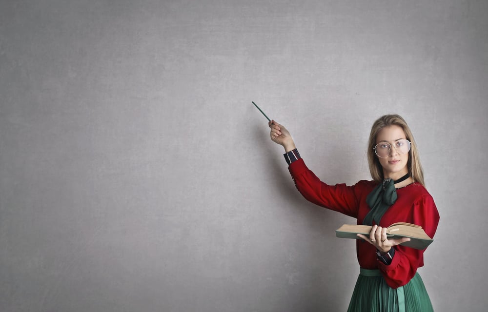 A person wearing big glasses and holding a book points to a blank blackboard.