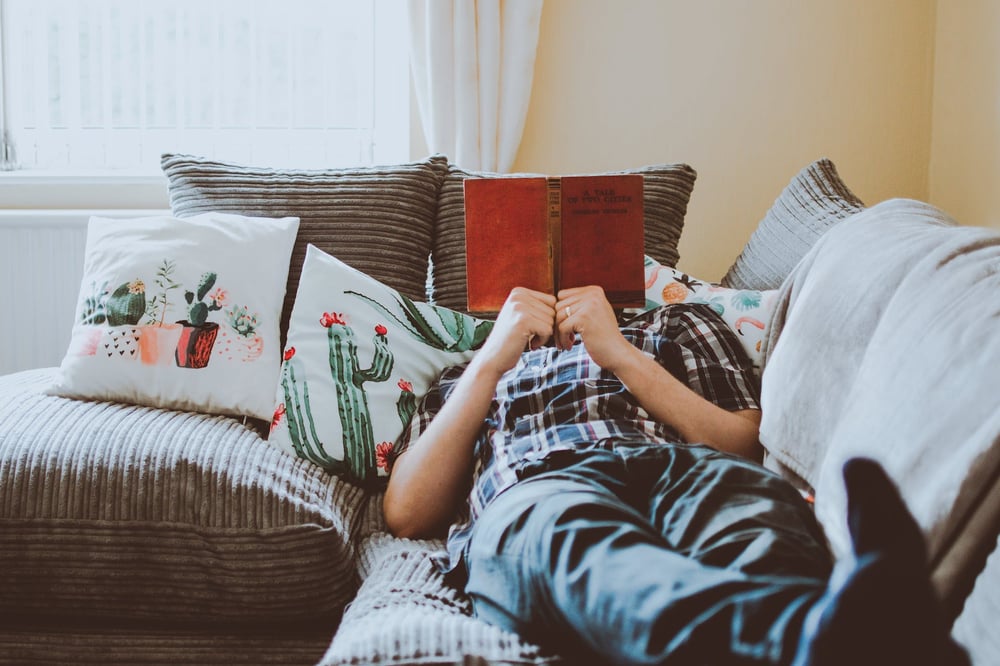 A person lying on a couch, their face obscured by the book they're reading.