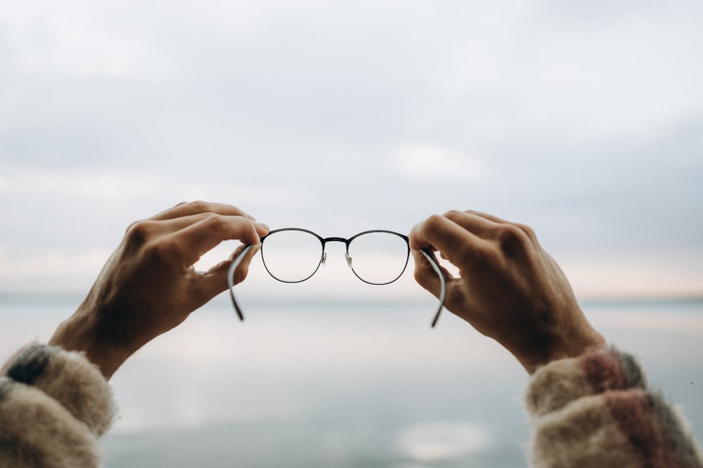 Hands hold a pair of glasses up, looking through to an ocean horizon.a