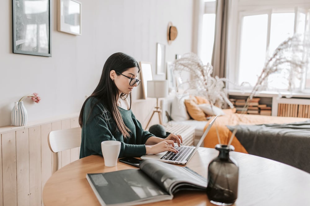 A writer sits at a table in a studio apartment, typing on a laptop.