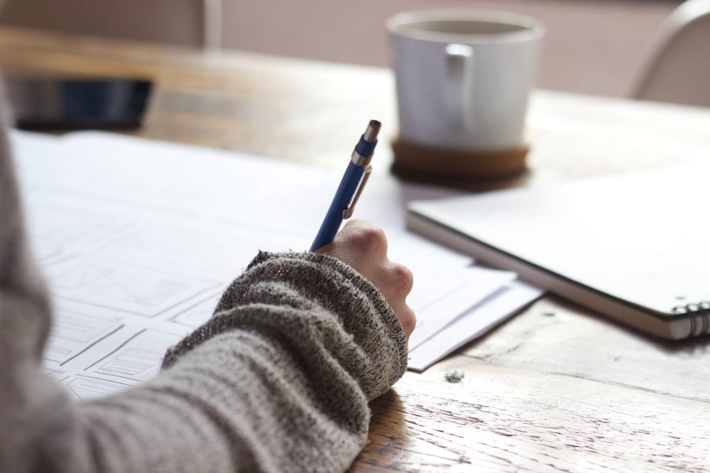 A hand writes on a piece of paper on a wooden desk.
