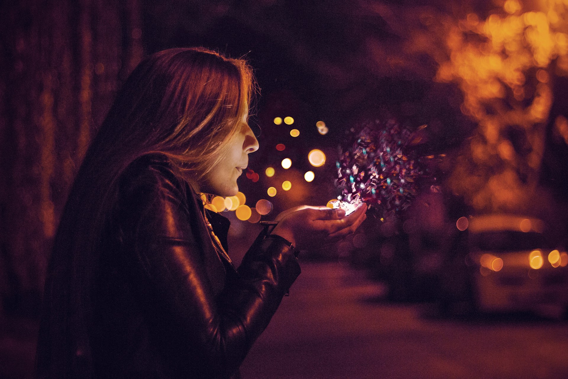A person in a leather jacket stands on a city street and blows glowing fairy dust out of their hands.