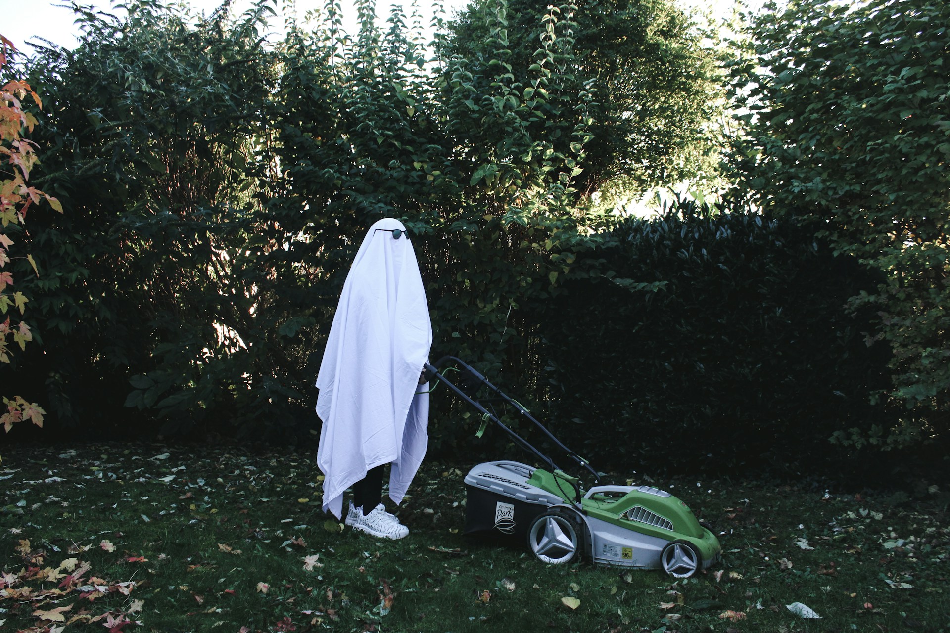 A person dressed in a sheet like a ghost and wearing sunglasses mows the lawn.