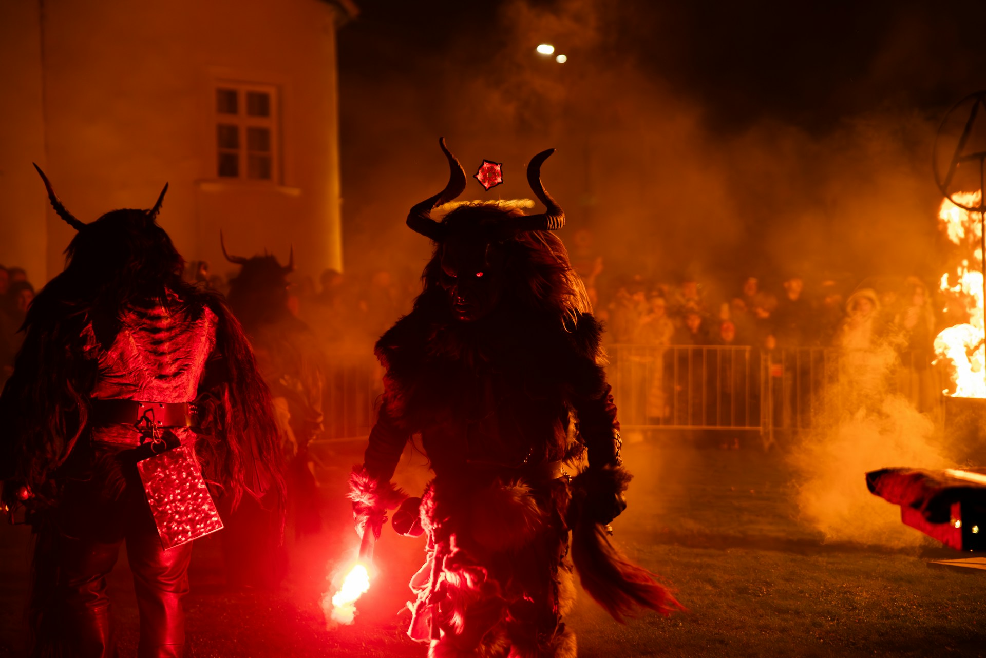 Red-eyed characters dressed in horns and fur walk around carrying small torches as a crowd looks on.