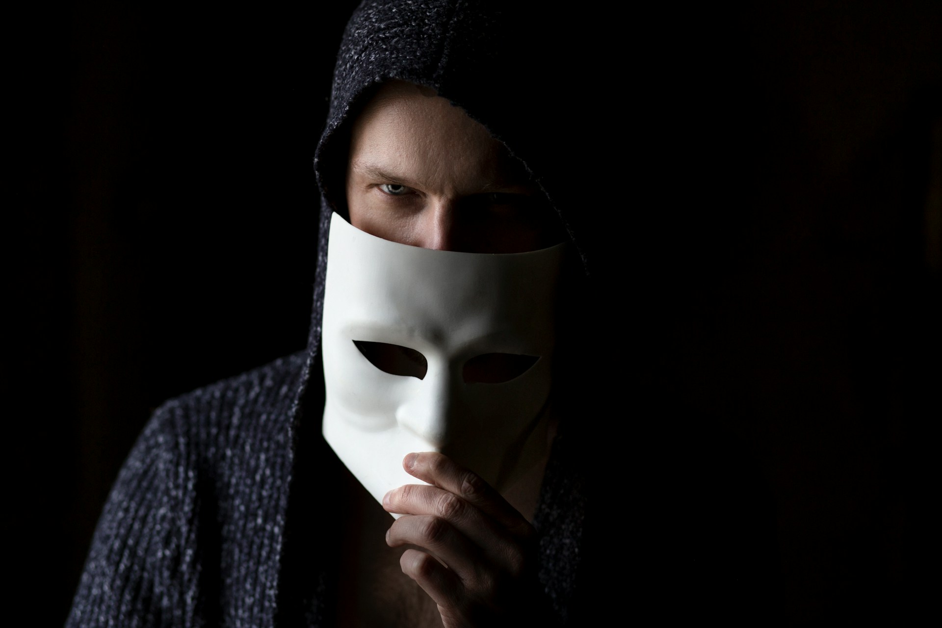 A person removes a plain, white mask halfway, revealing sinister-looking eyes.