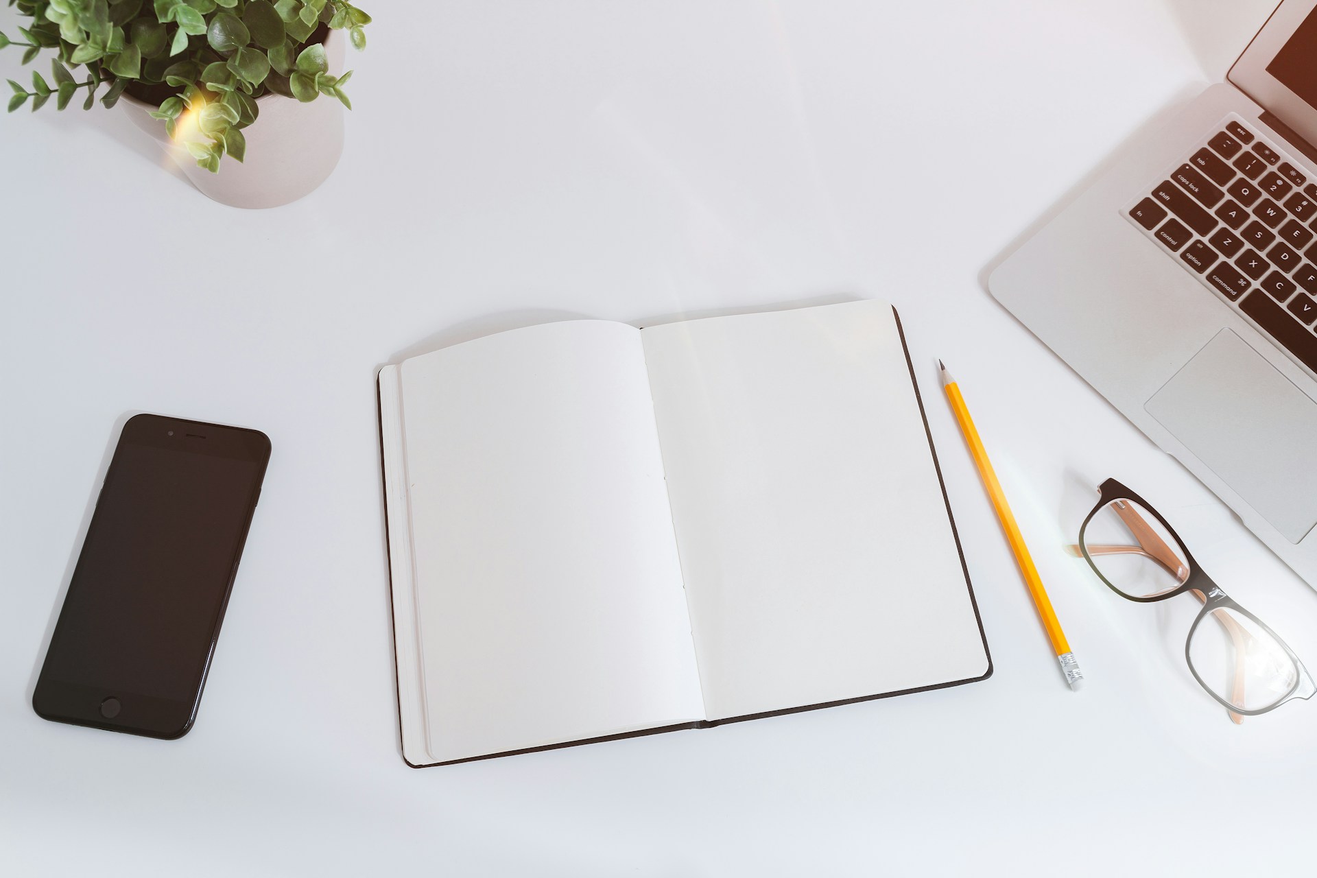 An open notebook sits on a white desk beside a smartphone, glasses, a pencil, and an open laptop.