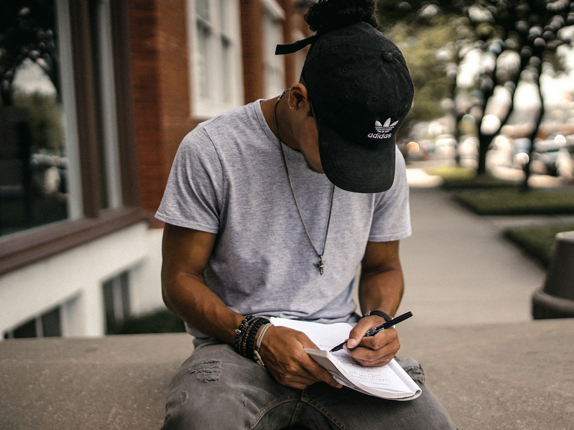 A person in a black Adidas hat sits on a concrete wall and writes in a notebook.