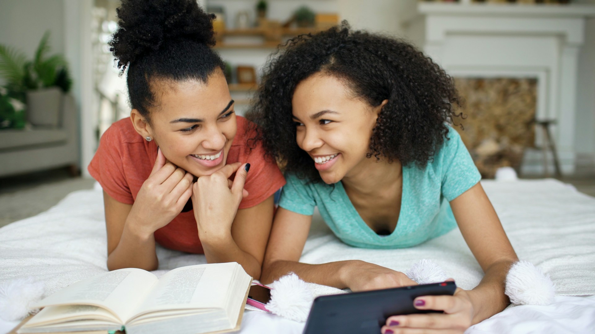Two young women lay side by side on a bed laughing over books together.