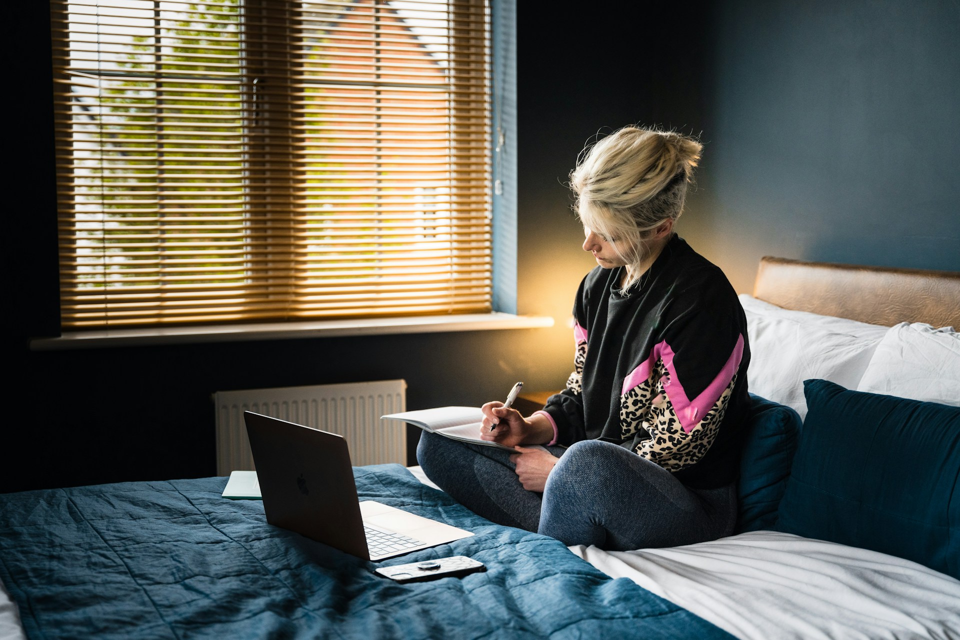 A person sits on a bed with a laptop, writing in a notebook.