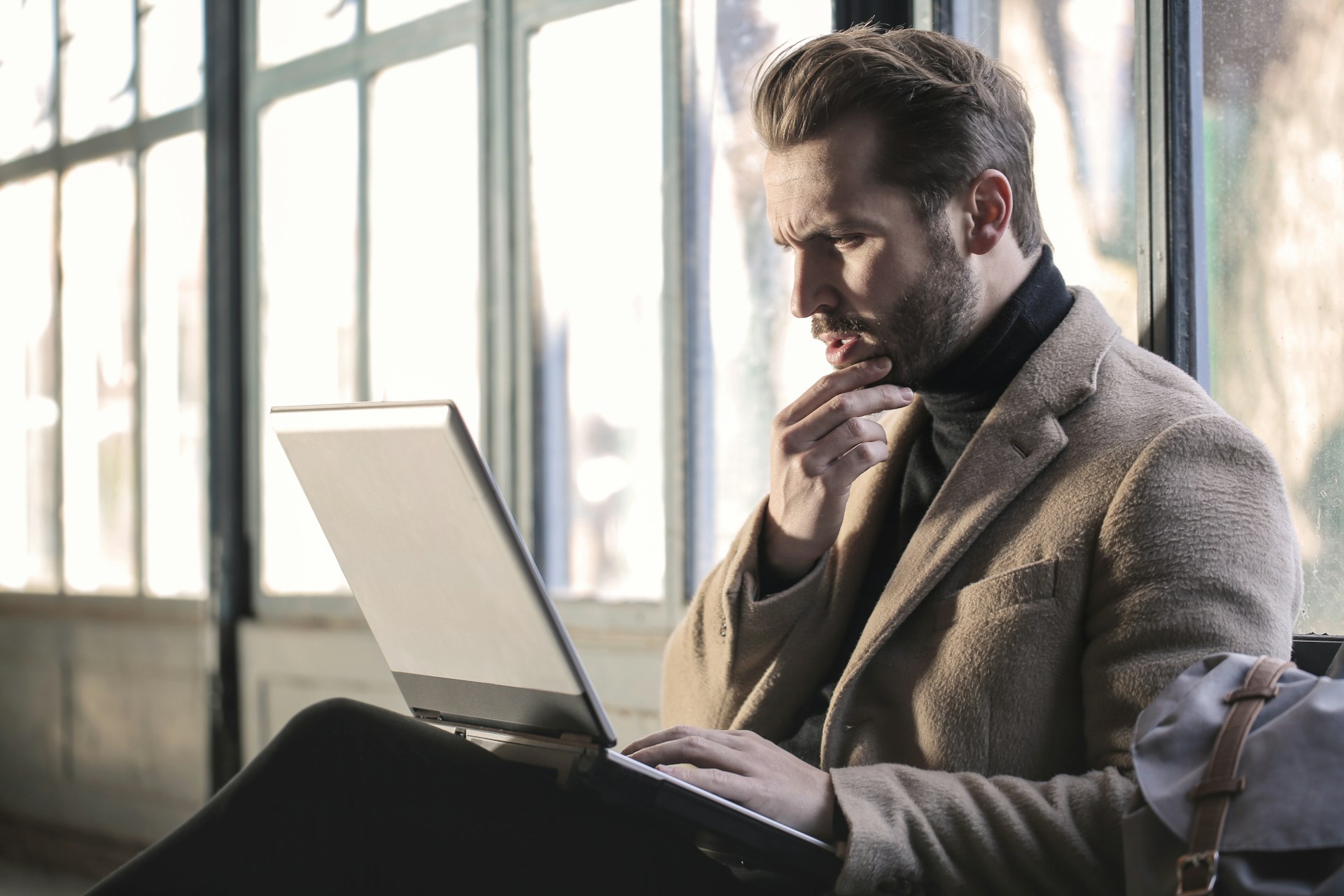 A person in a turtleneck and sport coat sits looking at a laptop screen with a confused expression on their face.