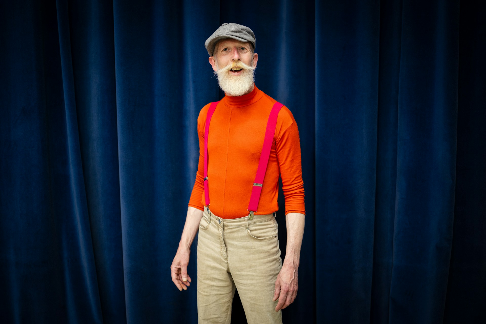A person with a red shirt, red suspenders, and a beard stands in front of a navy curtain, smiling.