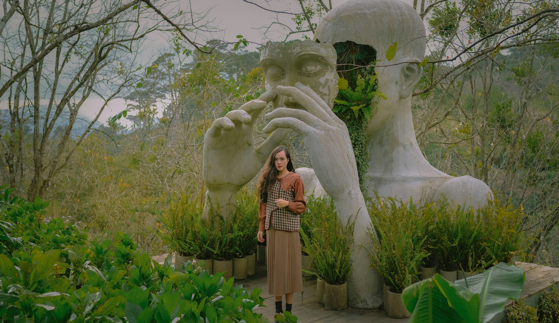 A person stands in a park in front a huge statue of a person whose face is removed to reveal plants.