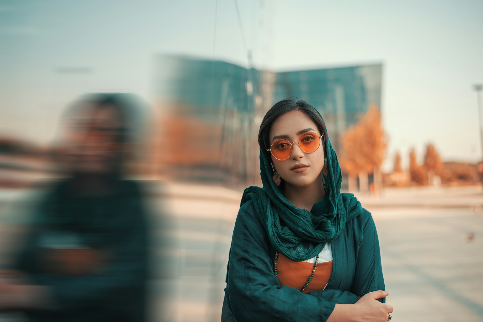 A person in a teal headscarf and orange glasses leans against a wall outside.