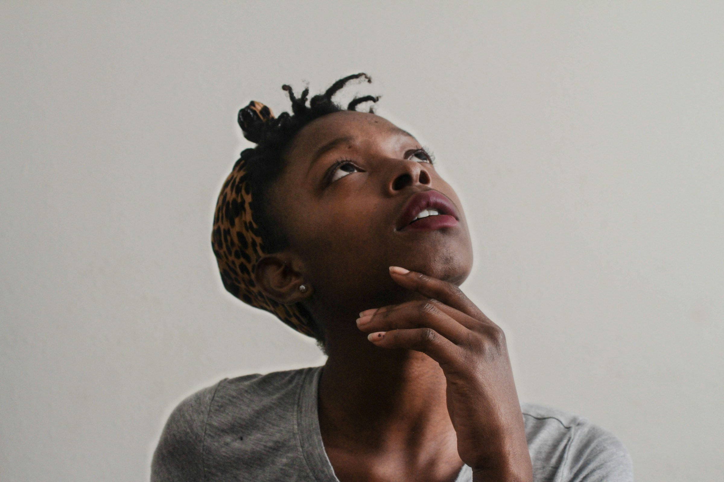 A person wearing a scarf around their hair looks upward and touches their chin as if thinking.