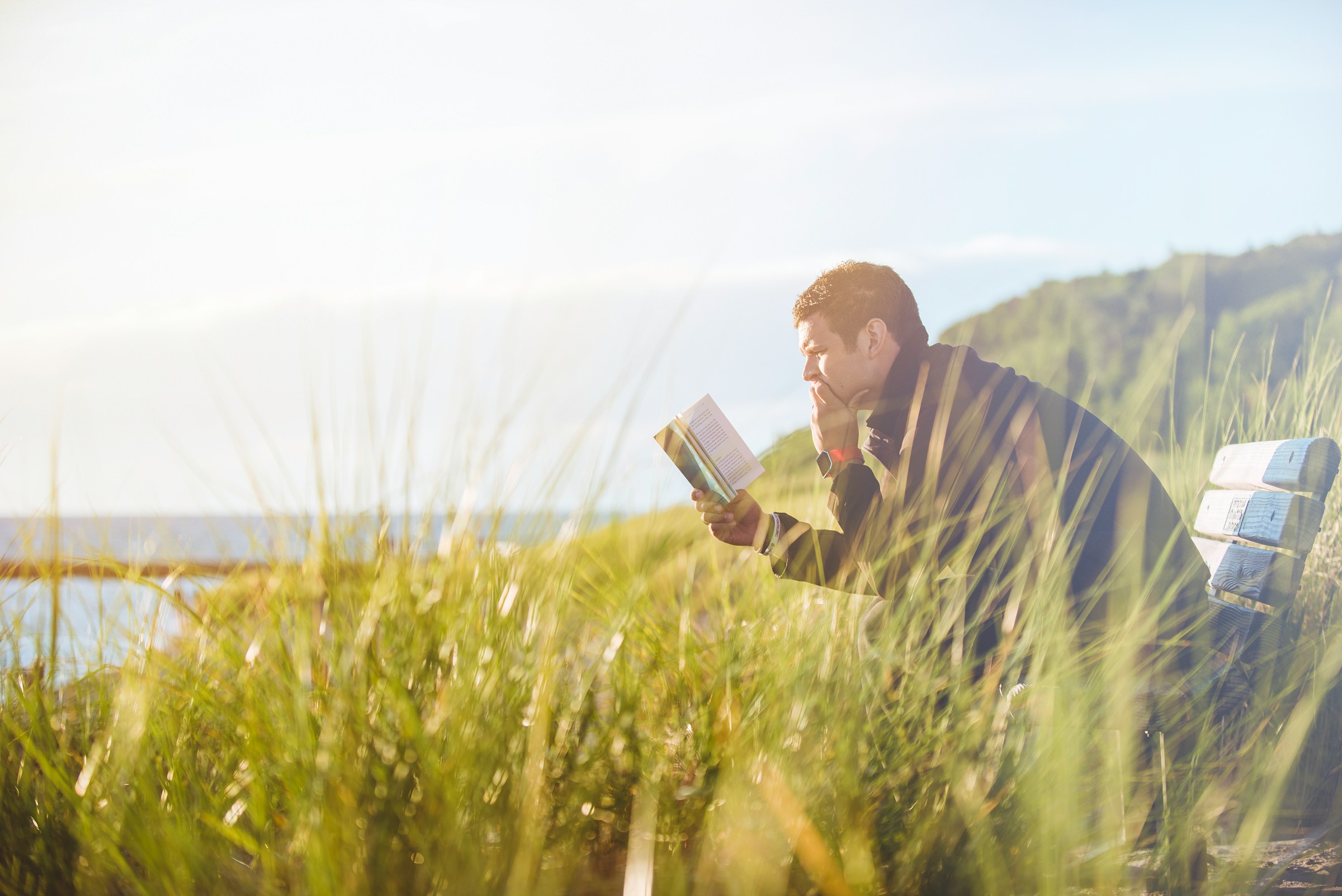 A person reads a book on a bench on a grassy hillside beside a body of water.