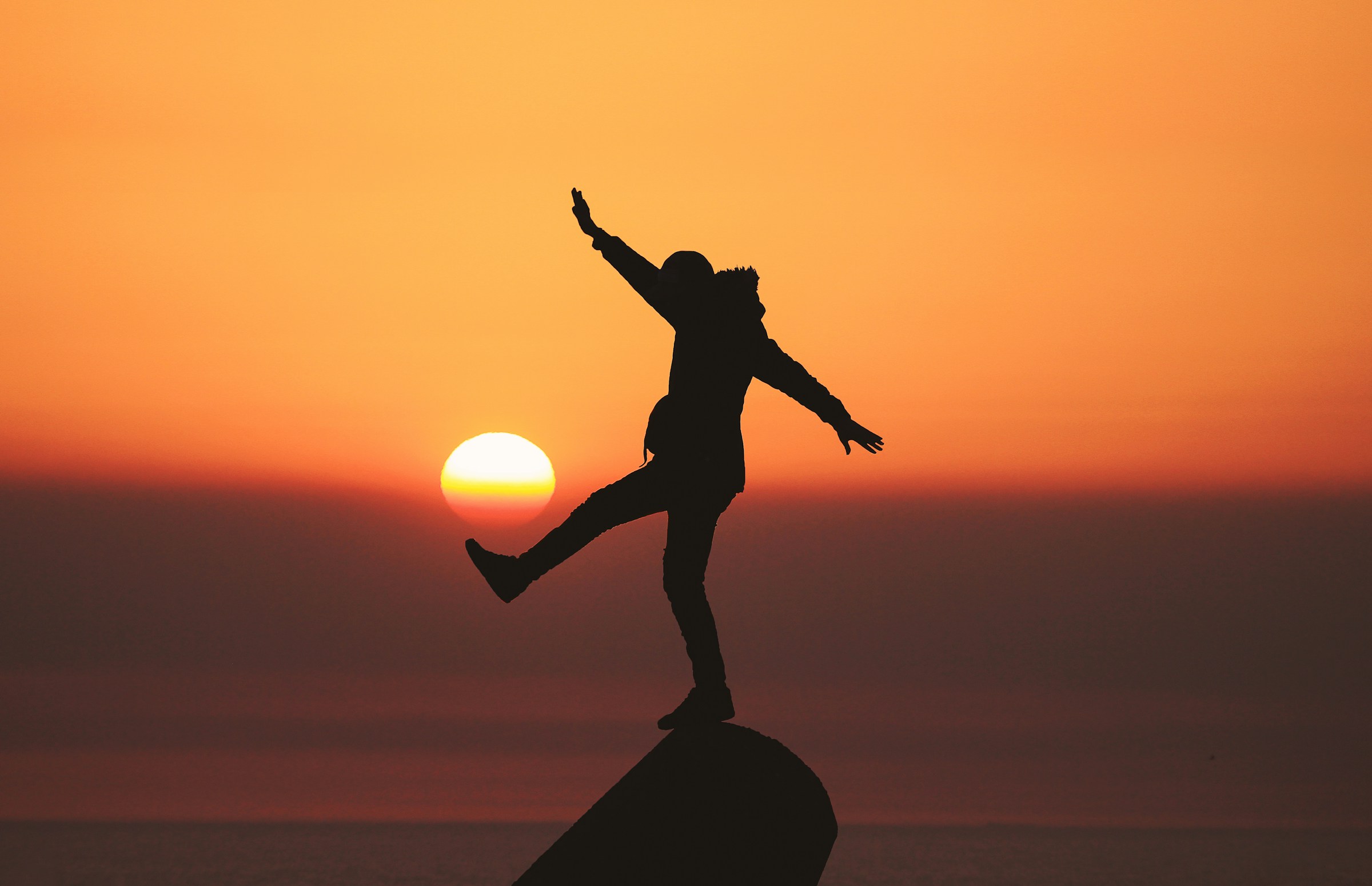 Silhouette of a person balancing on a rock with their arms in the air as the sun sits low in an orange sky behind them.