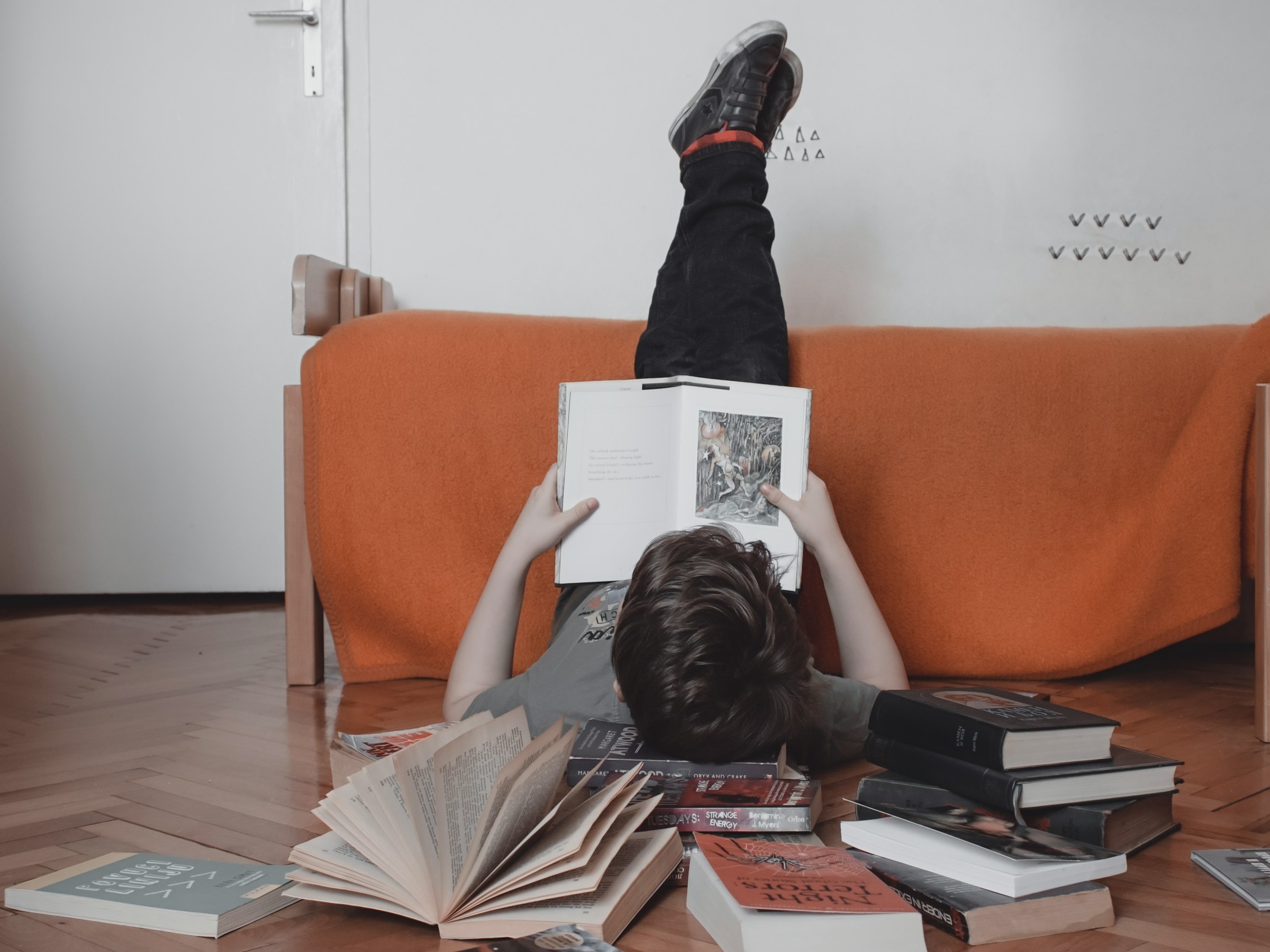A kid lies on the floor on their back with their legs propped up on a bed, reading a book.