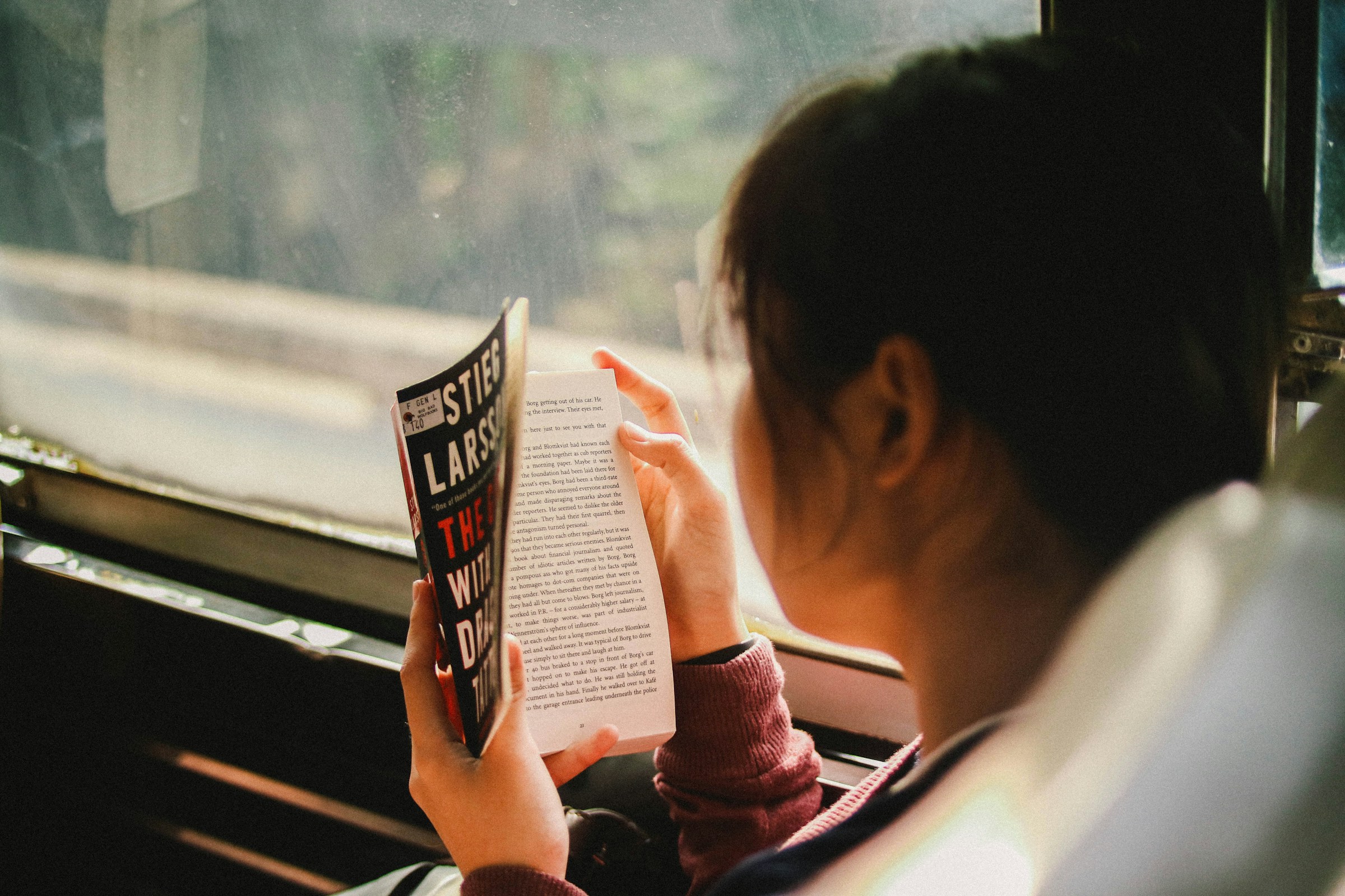 A young person sits on a bus or train beside a window, reading a book.