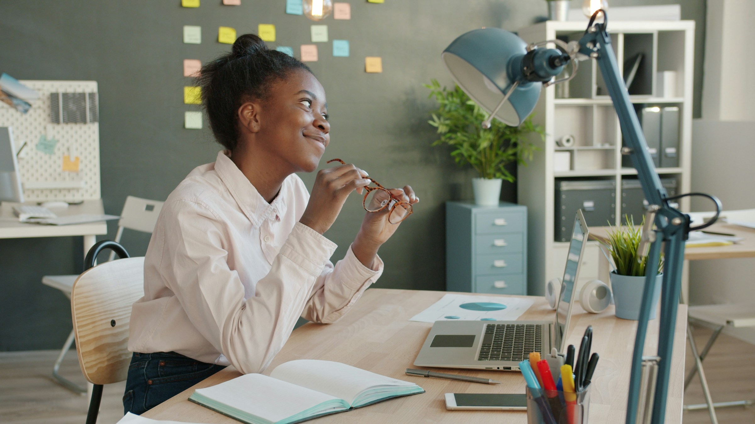 A person sits at a desk holding their glasses and smiling at something or someone across the room.