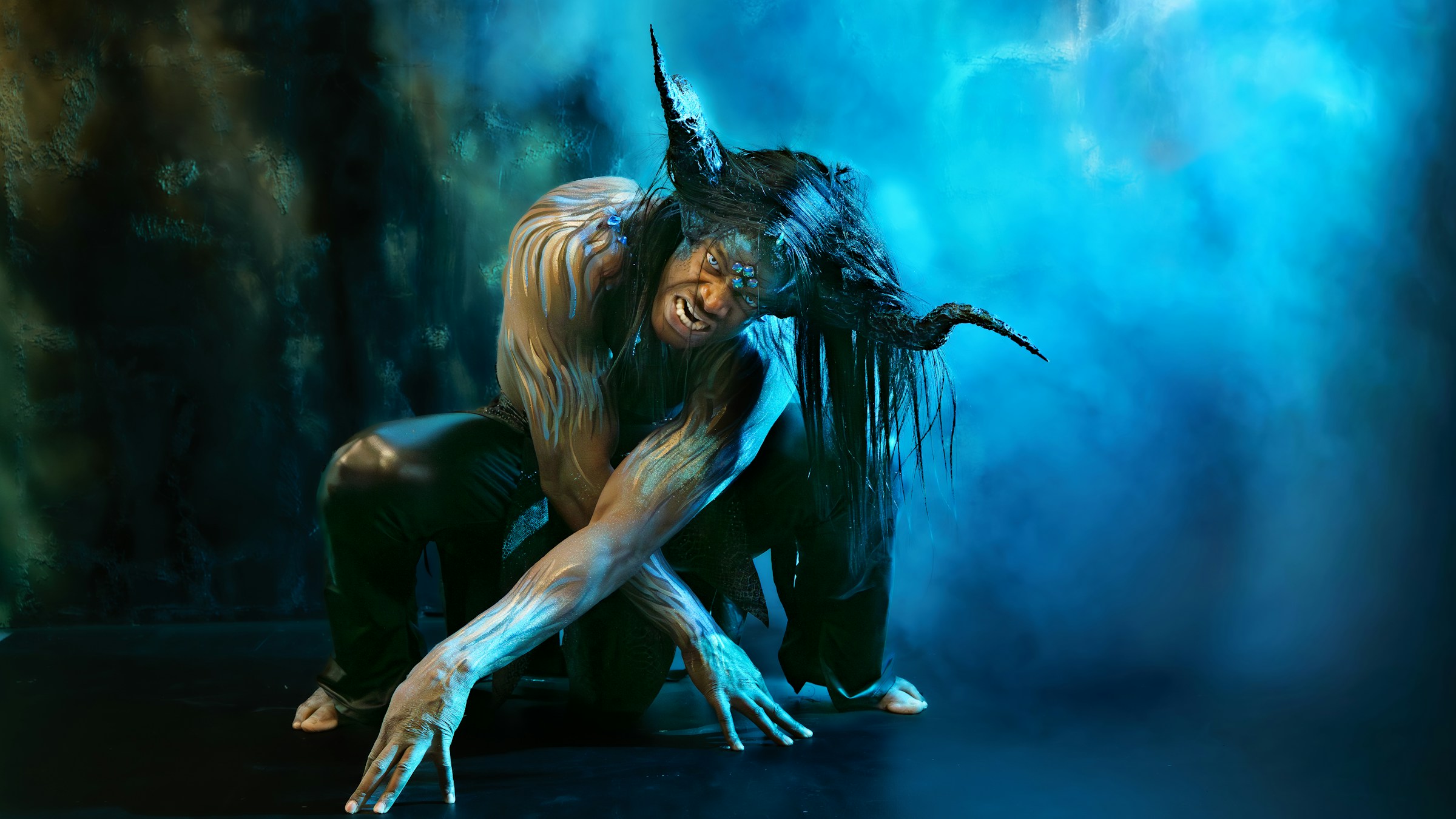 A half-human with long greasy hair and curved horns crouches in an unnatural position and scowls.
