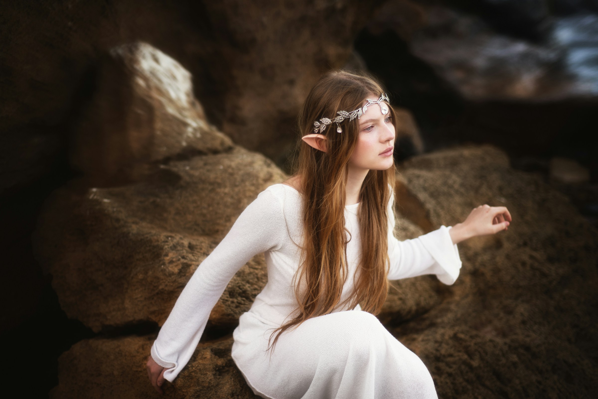 An elf with long hair and a long, white dress sits on large rocks.