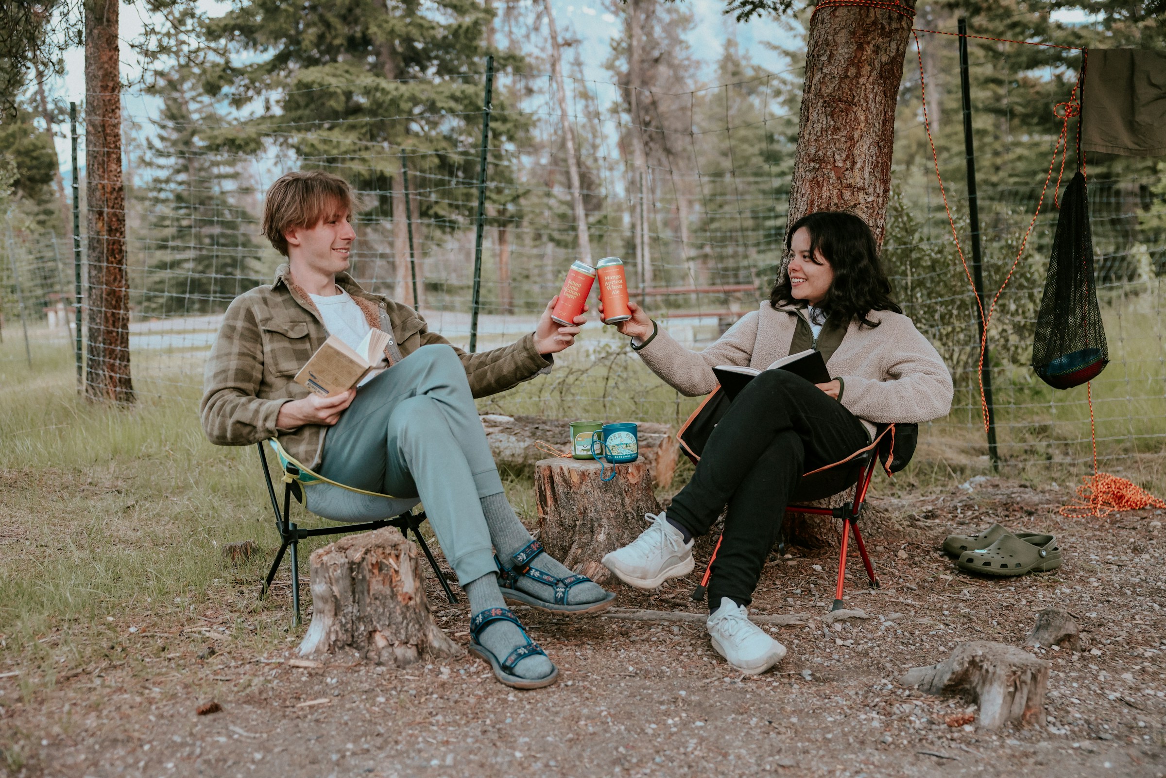 Two people sit outside in camp chairs, holding books and clinking beverage cans.